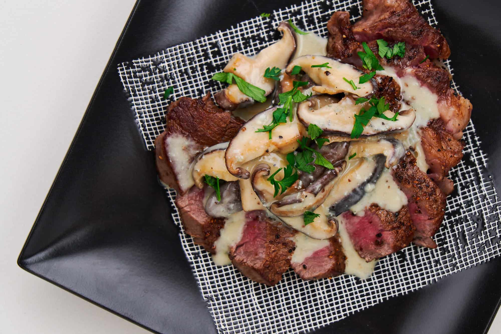 Juicy steak slices covered with shiitake and Gorgonzola sauce, garnished with fresh parsley on a modern black plate.