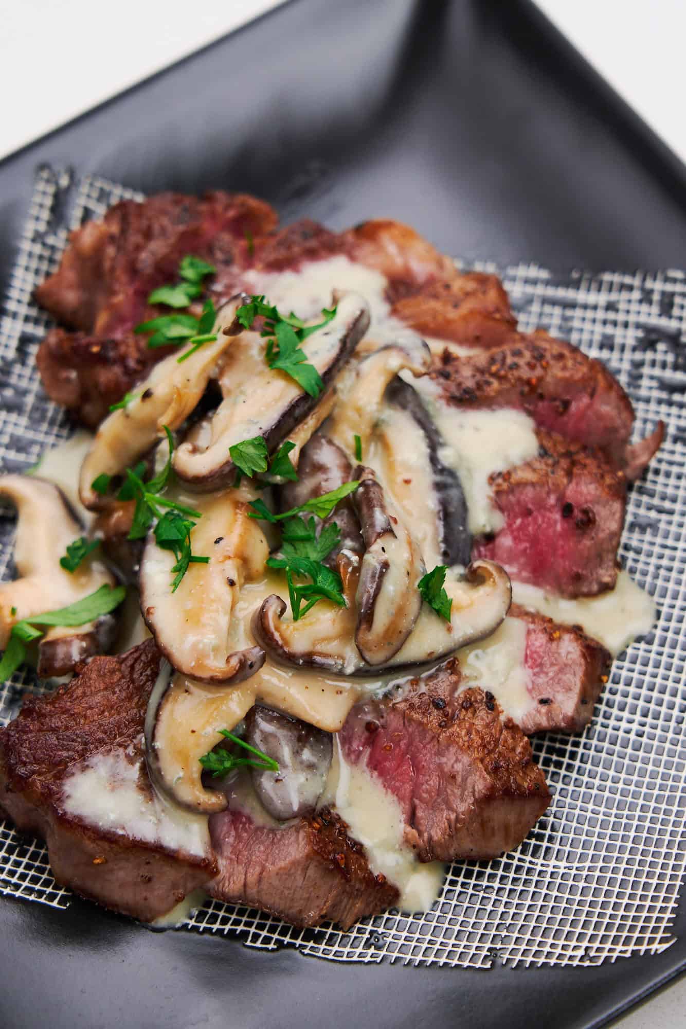 Close-up of steak with creamy shiitake Gorgonzola sauce and parsley garnish, served on a square black dish.