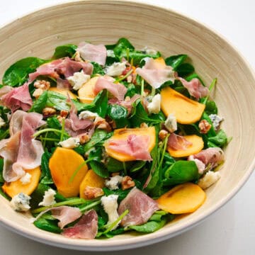 Close, horizontal beauty shot of the persimmon salad highlighting glossy greens, creamy blue cheese, toasted walnuts, and silky pieces of cured ham.