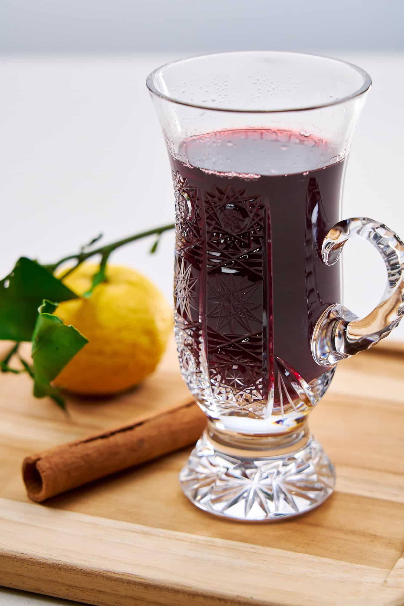 Footed crystal mug filled with Japanese mulled wine (glühwein) on a wooden board beside a whole yuzu and cinnamon stick.